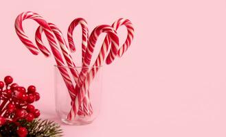 Studio shot of beautiful minimalistic simple composition with Christmas lollipops in transparent glass and snowy branch of pine with red berries, holly, on pink background with copy space for ad photo