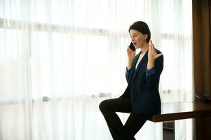 mujer de negocios, hembra empleado, joven empresario jura, argumenta, gestos con su manos mientras hablando en el teléfono, sentado a el borde de un mesa por ventana de un hotel habitación durante un negocio viaje foto