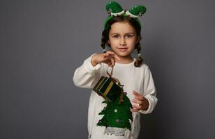 Navidad publicidad concepto con adorable niño niña en duende aro y blanco suéter con Navidad árbol participación Navidad regalo caja , sonriente linda mirando a el cámara. Copiar espacio en gris antecedentes foto
