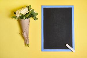 Flat lay composition of a delicate bouquet of autumn asters flowers in craft wrapping paper tied with a rope and blank empty chalkboard with space for text isolated on yellow background photo