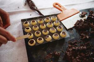 Confectioner hands putting chocolate filling into spherical molds from white chocolate and preparing chocolate truffles at home kitchen photo