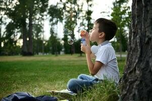 Portrait of an adorable cheerful beautiful cute elementary aged school boy enjoying his recreation between classes, blowing soap bubbles, sitting on the green grass of urban city park photo