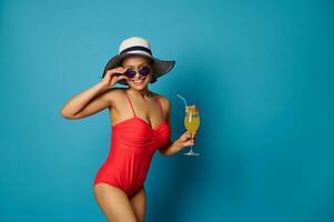 Sporty woman in a red swimsuit and summer hat holding a glass of juice and looking at camera through sunglasses photo