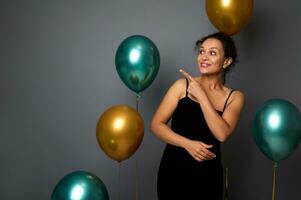 Beautiful Hispanic woman in black velvet evening dress, smiles looking to the side pointing with index finger on copy space on gray background with air balloons. Birthday, Christmas, New Year concept photo