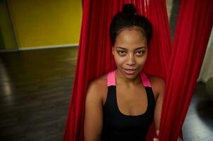 Close-up portrait of African woman with slim body in a black activewear practicing aerial fly yoga on a red hammock photo