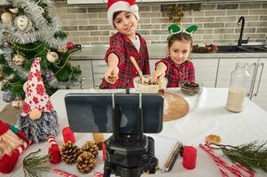 dos pequeño bloggers cocineros, adorable europeo niños, grabación un vídeo Blog mientras Cocinando juntos, demostración Pasas en un de madera cuchara en su manos a un teléfono inteligente cámara montado en un trípode. foto
