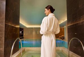 Rear view of a dark-haired beautiful woman in white terry robe standing by the pool and jacuzzi in luxury health spa enjoying relaxation and self-care. Beauty treatment, relaxation, body care concept photo