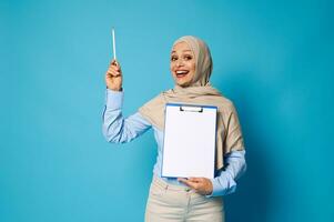 Smiling Arab woman holding a clipboard with a white blank sheet of paper and points a pen on blue background with space for text photo