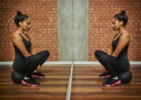 retrato de un hermosa deportivo atleta africano mujer en negro ropa de deporte mirando a su espejo reflexión, sentado en un medicina aptitud pelota en contra rojo ladrillos pared antecedentes de un gimnasio clase foto