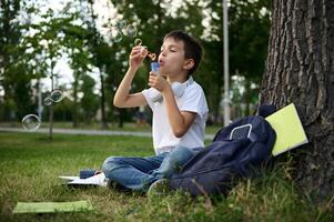 A schoolboy of elementary grades resting in the park sitting on the green grass after school, blowing bubbles. School bag with workbooks and school supplies lying down on the grass photo