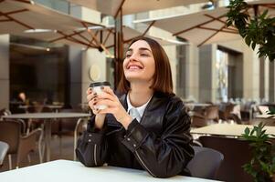 Attractive young woman with closed eyes of pleasure, enjoying coffee break, resting in outside cafe, holding a takeaway paper cup with hot drink, smiling looking up. Freedom, freelancer, lifestyle. photo