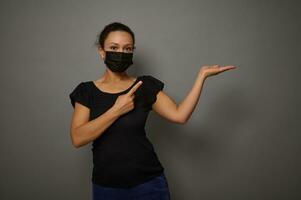 Black Friday concept. Beautiful brunette mixed race woman in black protective medical mask holds an imaginary copy space on and points with index finger on it, smiles looking at camera photo