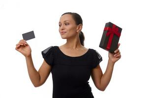 Beautiful mixed race Hispanic woman looks at plastic credit or discount card in her hand and holds a black gift box with red ribbon in her other hand . Concept of Black Friday with copy space photo