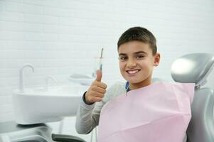 Cheerful confident brave handsome elementary aged child, school boy shows thumb up and smiles with beautiful toothy smile looking at camera while sitting in dentist's chair after dental treatment photo