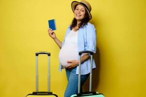 Happy pregnant woman putting her hand on her belly, smiling at camera, posing with boarding pass over yellow background photo