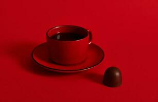 Closeup of a red ceramic cup of coffee on a saucer and a chocolate praline on red background with copy space . Concept of chocolate day photo