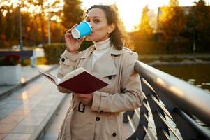 Serene African woman in beige cozy trench coat drinks coffee from takeaway paper cup, reads a hand book and enjoys her day off outside in beautiful autumn park with lake at sunset photo