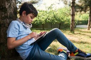 Hispano adolescente chico disfrutando su creativo pasatiempo, dibujo bocetos en un digital tableta, utilizando un aguja bolígrafo, sentado en parque foto