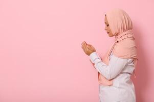 Side portrait of a serene Muslim Arab woman in pink hijab and strict outfit praying, performing namaz, isolated on colored background with space for text photo