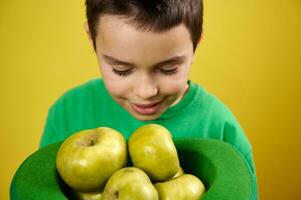 Little boy enjoys the scent of green apples in a green Irish cap. Portrait on yellow background. Saint Patrick's Day photo
