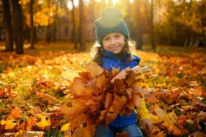 hermosa 4 4 años antiguo pequeño niña sentado en un dorado arce parque entre caído hojas a puesta de sol y sonrisas lindamente, mirando a el cámara, participación un recogido seco ramo de flores de otoño hojas en su manos foto