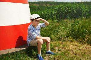 Adorable child boy sitting on the background of a bright lighthouse and looking into the distance. photo