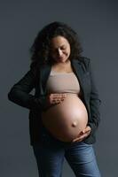 elegante multiétnico embarazada mujer sonrisas mientras suavemente acariciando su grande barriga en el embarazo 36 semana, sensación bebé patadas foto