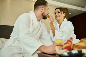 Happy loving married couple, newlyweds sitting on bed after waking up and enjoying delicious breakfast for two in luxury hotel bedroom while spending their honeymoon. Tenderness, love, affection photo