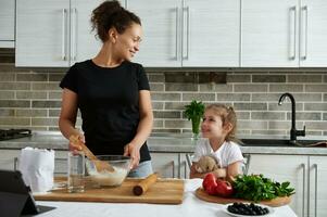 mezclado carrera mujer, mirando a su linda bebé niña mientras amasadura masa. madre y hija Cocinando juntos en el cocina foto