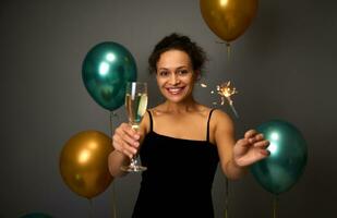 Happy Latin American woman holds flute of champagne and sparklers, smiles with beautiful toothy smile poses against gray wall background with inflated golden and green air balloons. Copy space for ad photo