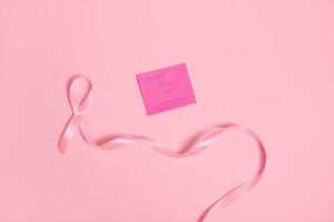 High angle view of a long pink ribbon with endless one end, and a paper note with lettering 1 October Breast Cancer Awareness , isolated on pink background with copy space. Flat lay photo