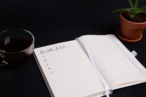 Opened notepad, organizer with list to do on blank white sheets, cup of coffee and potted succulent in clay pot, on black background with copy space. Business, Organization, Time Management concept. photo