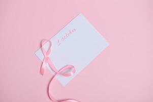 Flat lay of a pink ribbon with an endless end lying on a white paper sheet with lttering 1st October, International symbol of Breast Cancer Awareness Month. Woman's healthe care and medical concept photo