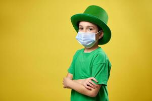 Side portrait of a boy in green Irish hat and protective medical mask poses to camera with arms crossed. Saint Patrick day photo