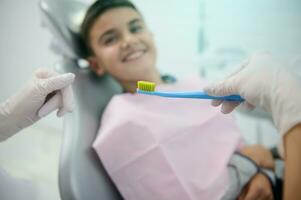 Soft focus on a toothbrush in the hand of a doctor dentist teaching an out of focus blurred smiling handsome boy sitting in dentist chair about oral care and dental everyday hygiene. Medical concept photo