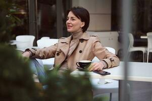 Toned photography, portrait of attractive beautiful young brunette woman in stylish casual wear smiling toothy smile looking away enjoying coffee break sitting at the table outdoor photo