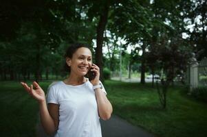 hermosa multiétnico joven mujer hablando en móvil teléfono mientras caminando a lo largo el callejón de un ciudad parque en un verano día foto