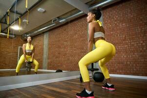 Beautiful young African woman with healthy slim body and flat belly in stylish bright yellow tracksuit squatting with dumbbells while working out in fitness studio. Body care, weight loss concept photo