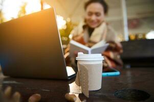 Takeaway cardboard paper cup with hot drink on table with laptop and blurred woman working remotely outdoors at beautiful autumn sunset. Autumn background with copy space photo