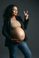 Attractive young pregnant woman posing bare belly on gray studio background, smiling and showing peace sign with fingers photo