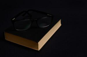 Monochrome composition of eyeglasses on a book in hard black cover, isolated over black background with copy space. Teacher's Day concept, Knowledge, literature ,reading, erudition photo