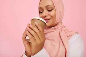 Arabic Muslim woman with covered head in pink hijab drinking hot drink, tea or coffee from disposable cardboard takeaway cup, standing three quarters against colored background, copy space. Close-up photo