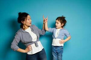 Young woman and her baby girl alike dressed looking at each other and pointing their index finger up. Blue background with copy space photo