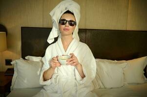 Beautiful woman in sunglasses and a white terry bathrobe with her head wrapped in a white bath towel, holding a cup of coffee and relaxing in the bedroom, sitting on the bed and looking at the camera photo