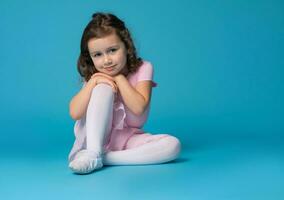 Adorable preschool girl child, ballet dancer, posing to the camera, sitting over blue background. photo