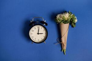 Flat lay composition of a beautiful bouquet of asters in craft paper and an alarm clock, lying on a blue background with space for text. Back to School and Teachers' Day Concepts photo