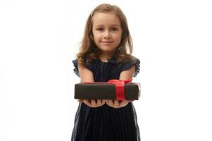 adorable pequeño niña en noche oscuro azul vestir participación un negro regalo caja con rojo cinta en su manos y demostración eso a cámara. aniversario, negro viernes concepto. aislado blanco fondo, Copiar espacio foto