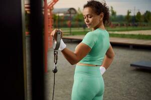 lado longitud de la cintura de un hermosa y atractivo juguetón mujer de Hispano etnia haciendo ejercicio en bíceps con estacionario al aire libre cable Transversal. determinado atleta trabajando fuera en campo deportivo foto