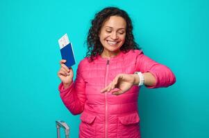 Happy curly dark haired young woman holding passport and boarding pass ticket smiling looking at watch, checking time, isolated on blue background with copy space. Travel, tourism, punctuality concept photo