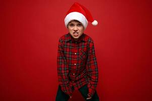 Funny preadolescent boy in plaid shirt and Santa Claus hat grimacing looking at camera posing against red colored background with copy space for Christmas and New Year ad photo